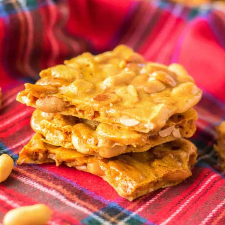 Pieces of peanut brittle stacked on top of one another with roasted peanuts sprinkled around them.