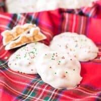 White chocolate polar bear paws candy with red and green sprinkles on top on a red plaid linen. One candy is cut in half to show caramel and peanuts inside.