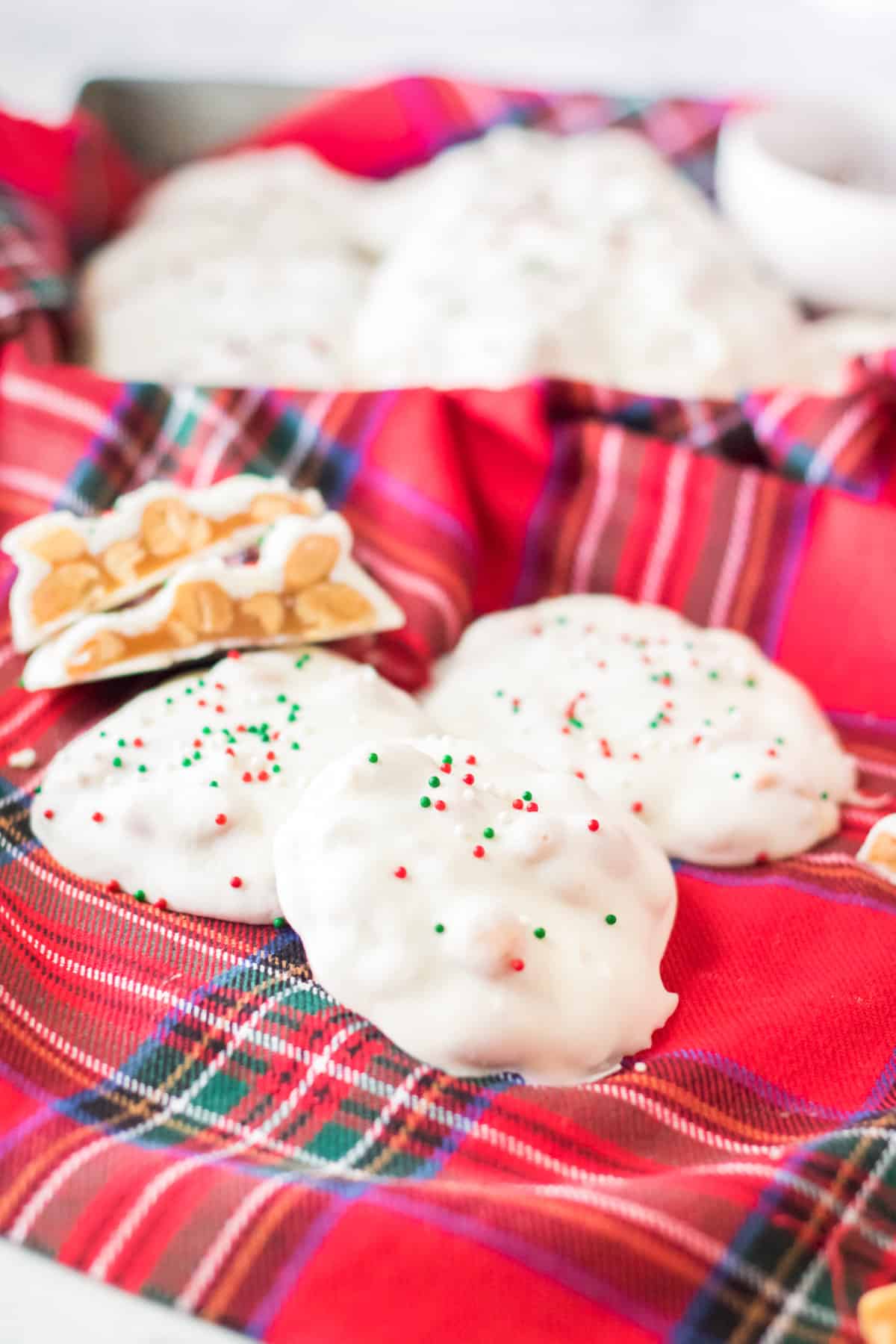 White chocolate polar bear paws candy with red and green sprinkles on top on a red plaid linen. One candy is cut in half to show caramel and peanuts inside.