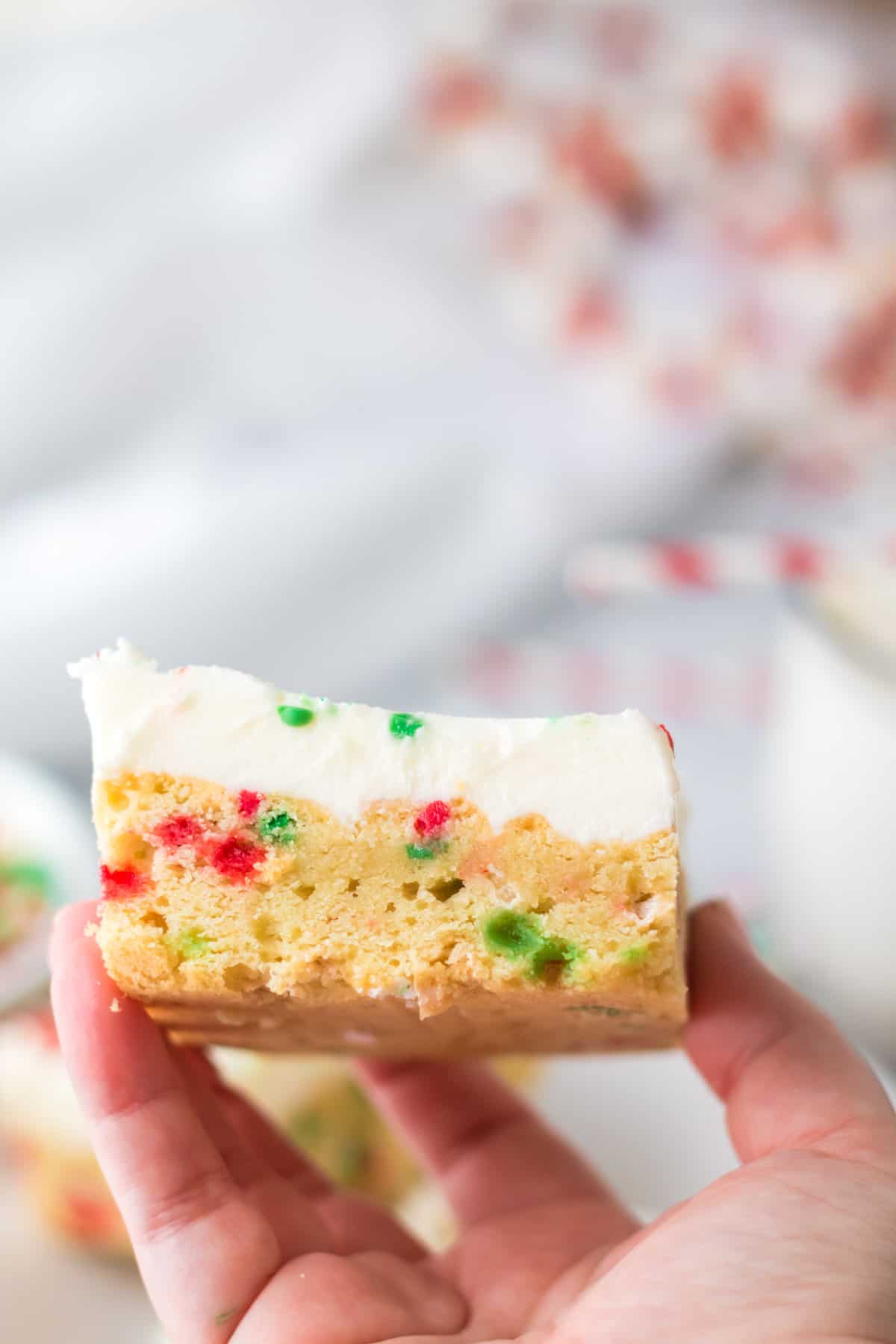 Hand holding thick and chewy frosted cookie bar with vanilla icing and christmas sprinkles.