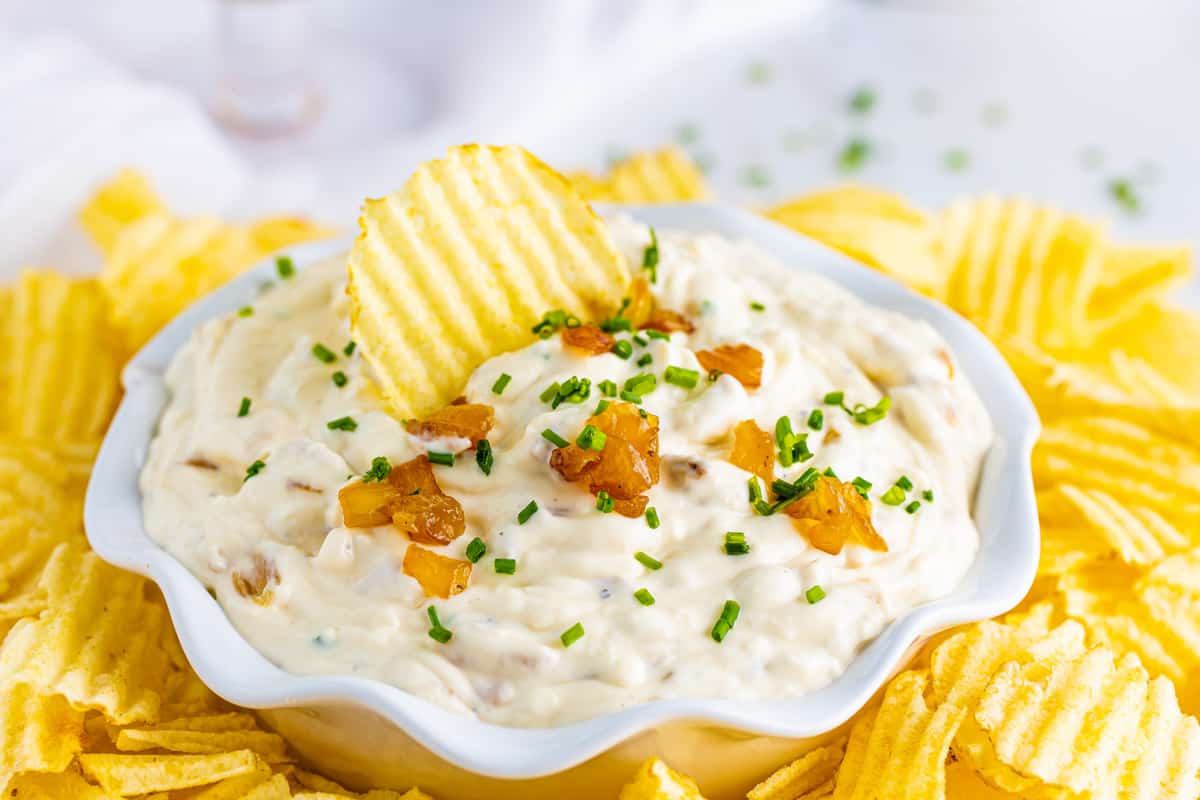 Bowl of homemade French onion dip served with wavy potato chips. One chip is sticking out of the top of the dip bowl.