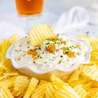 Homemade French onion dip served with wavy potato chips. One chip is sticking out of the top of the dip bowl.