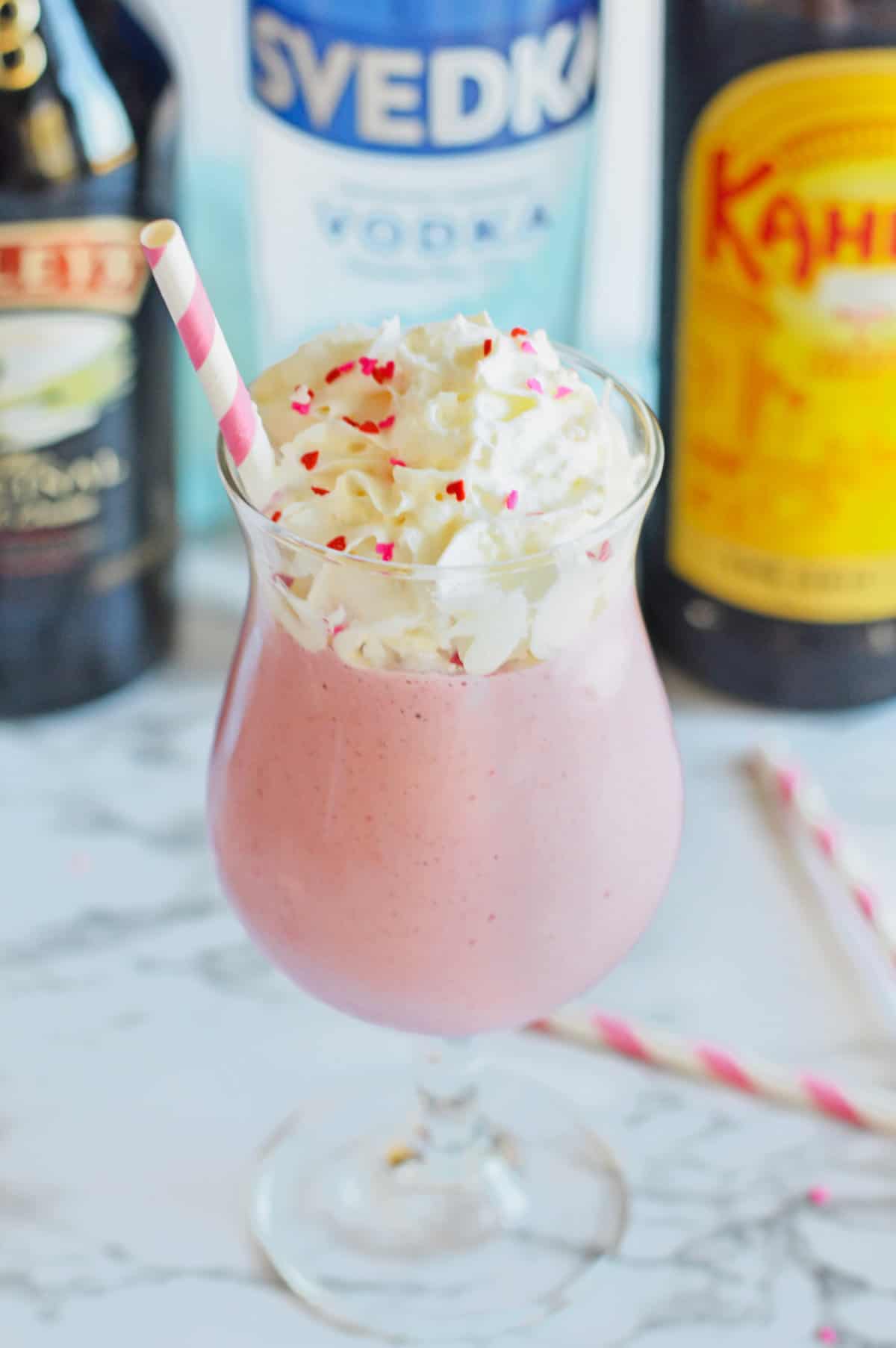 Pink strawberry mudslide in a glass with Kahula, vodka, and Baileys bottles behind it.