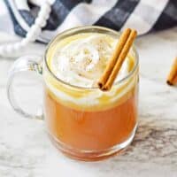 Hot buttered rum cocktail topped with whipped cream and cinnamon stick in a clear glass mug.