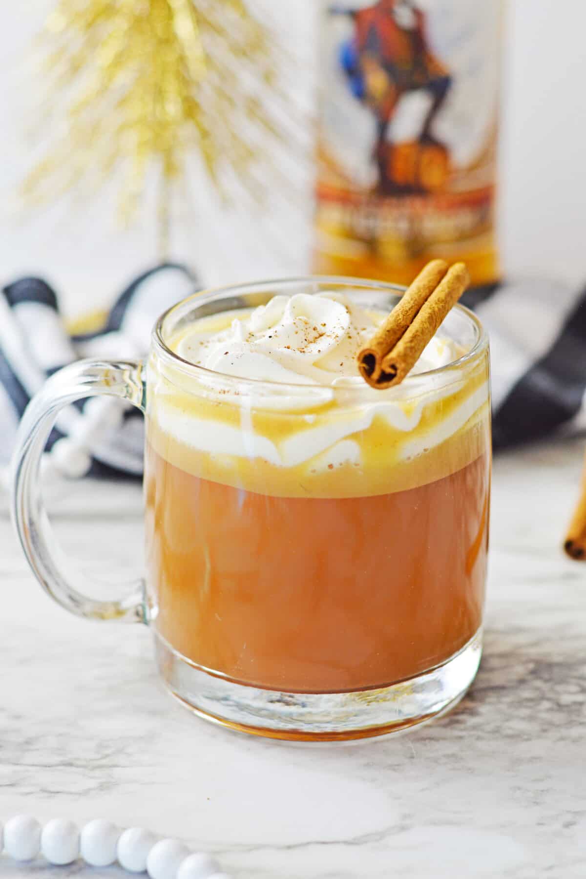 Hot buttered rum cocktail topped with whipped cream and a cinnamon stick in a glass mug with a bottle o spiced rum in the background.