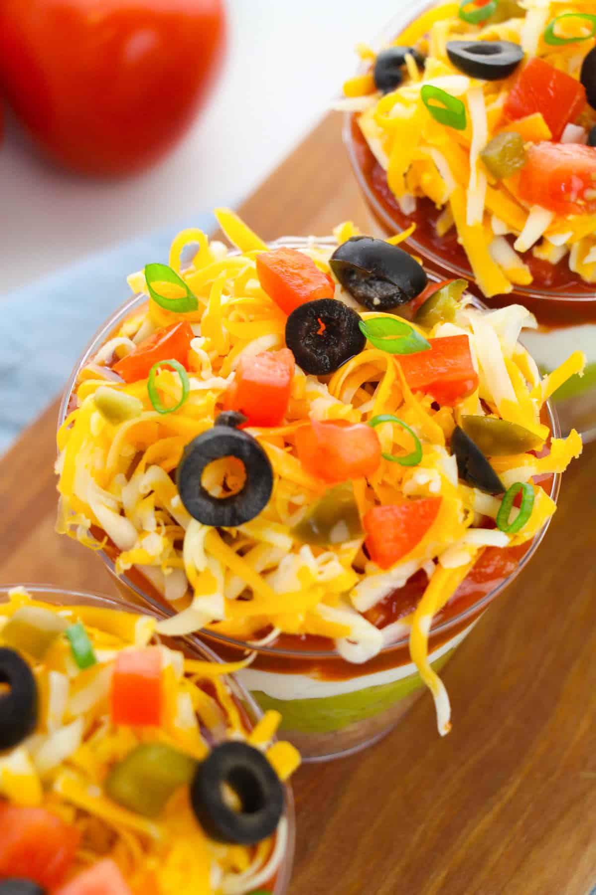 Top-down image of 7 layer dip in individual cups, showing the shredded cheese, tomato, olive, green onion, and diced jalapeno toppings.