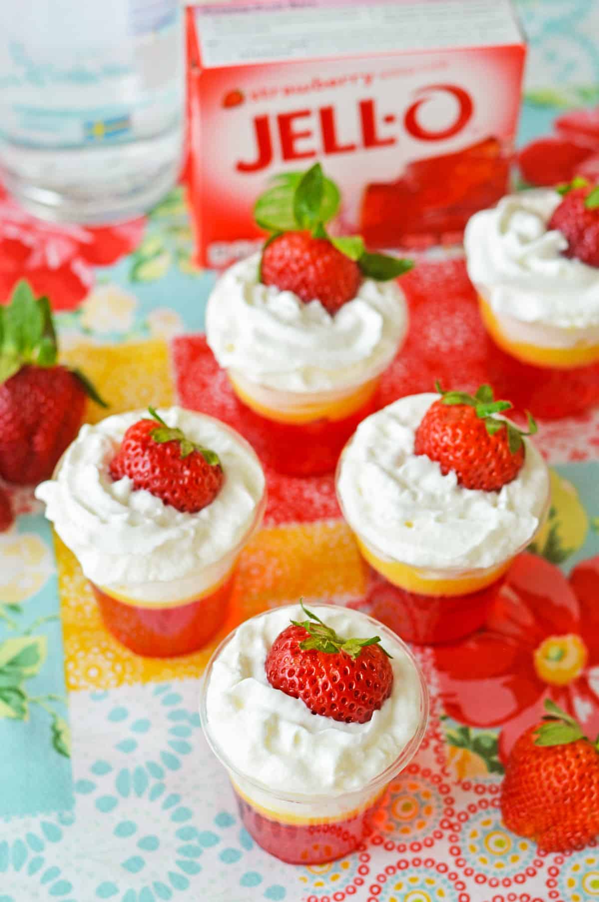 Five strawberry and cream jello shots with whipped cream and fresh strawberries on a patterned table cloth with a box of strawberry jello mix.