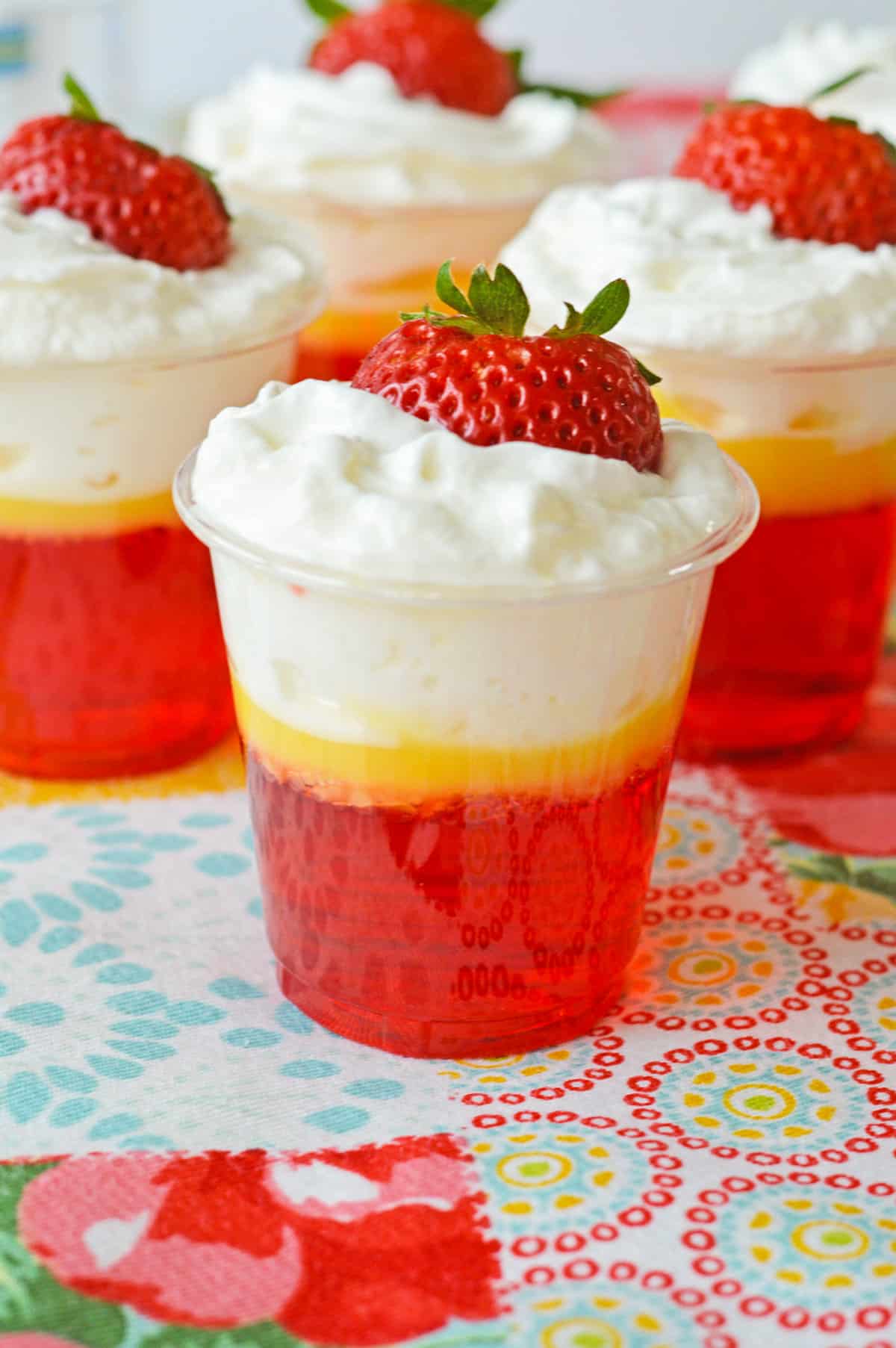 Strawberries and cream jello shots with 3 distinct layers: Strawberry jello, vanilla pudding, and whipped cream and topped with a fresh strawberry.