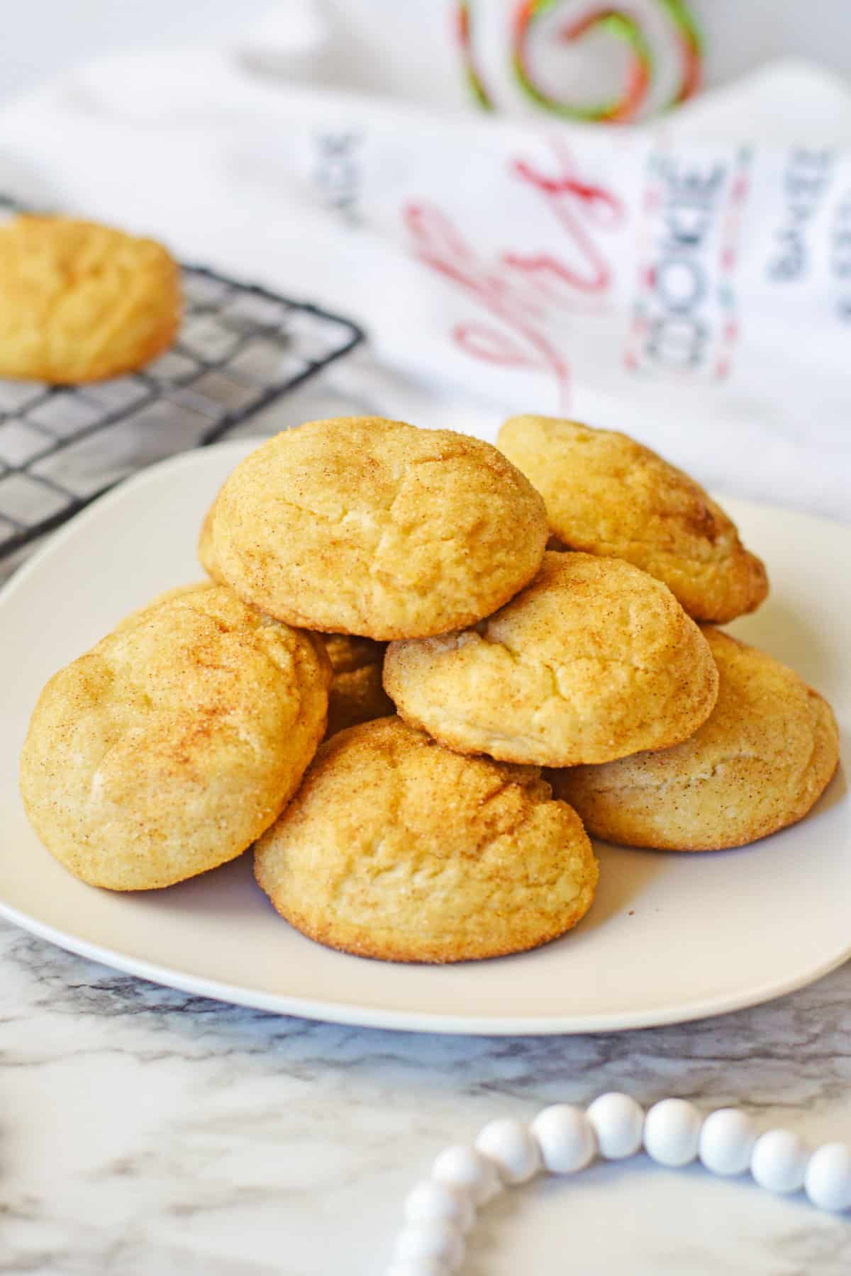 Snickerdoodle cake mix cookies stacked in a pile on a white plate with a cooling rack with more snickerdoodles behind them.