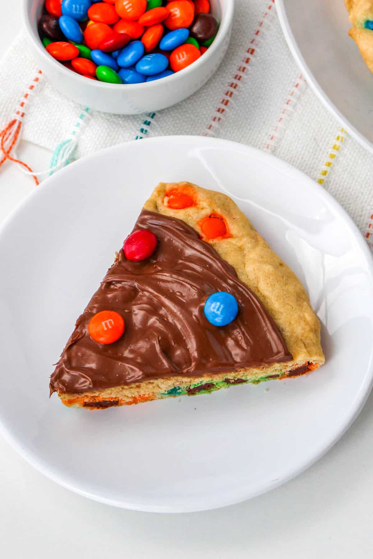 Slice of cookie pizza on white plate with bowl of M&Ms in background.