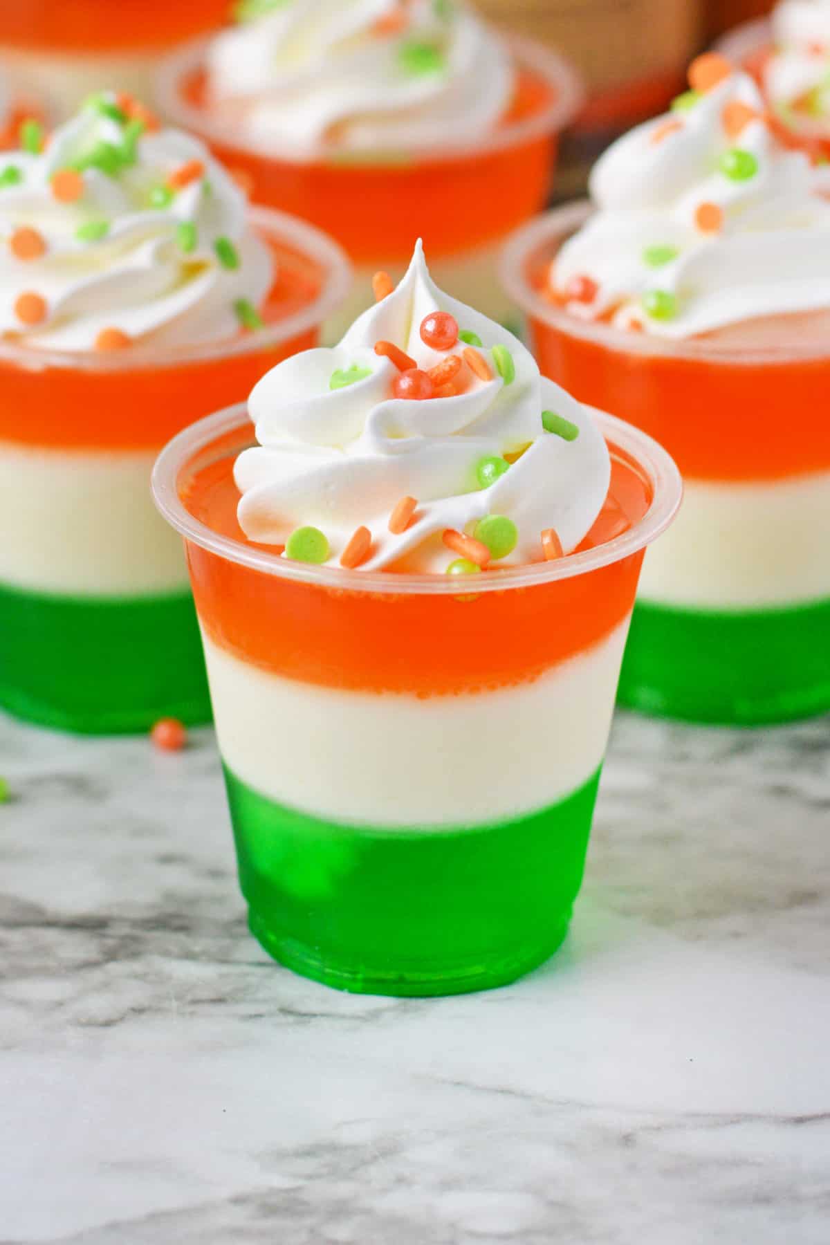 St. Patrick's Day jello shots made with a green bottom layer, white middle layer, and orange top layer, and whipped cream and festive sprinkles.