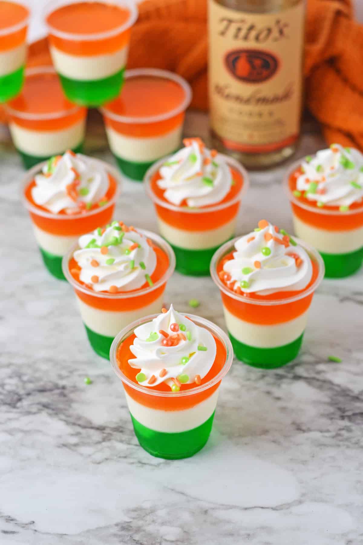 Whipped cream and sprinkle topped Irish flag jello shots with bottle of Tito's vodka behind them.