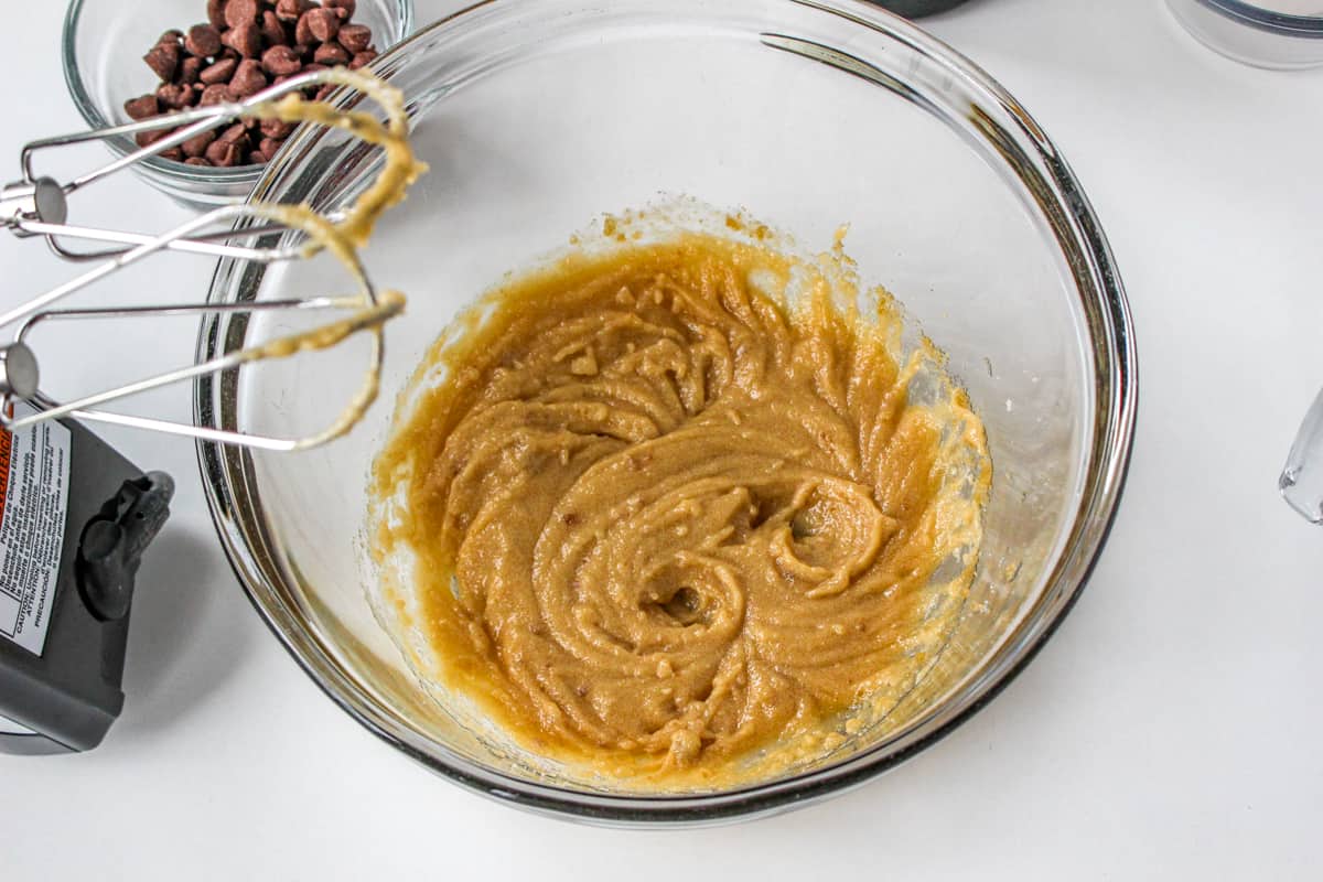 Melted butter combined with sugars in mixing bowl. An electric hand mixer is besides the bowl.