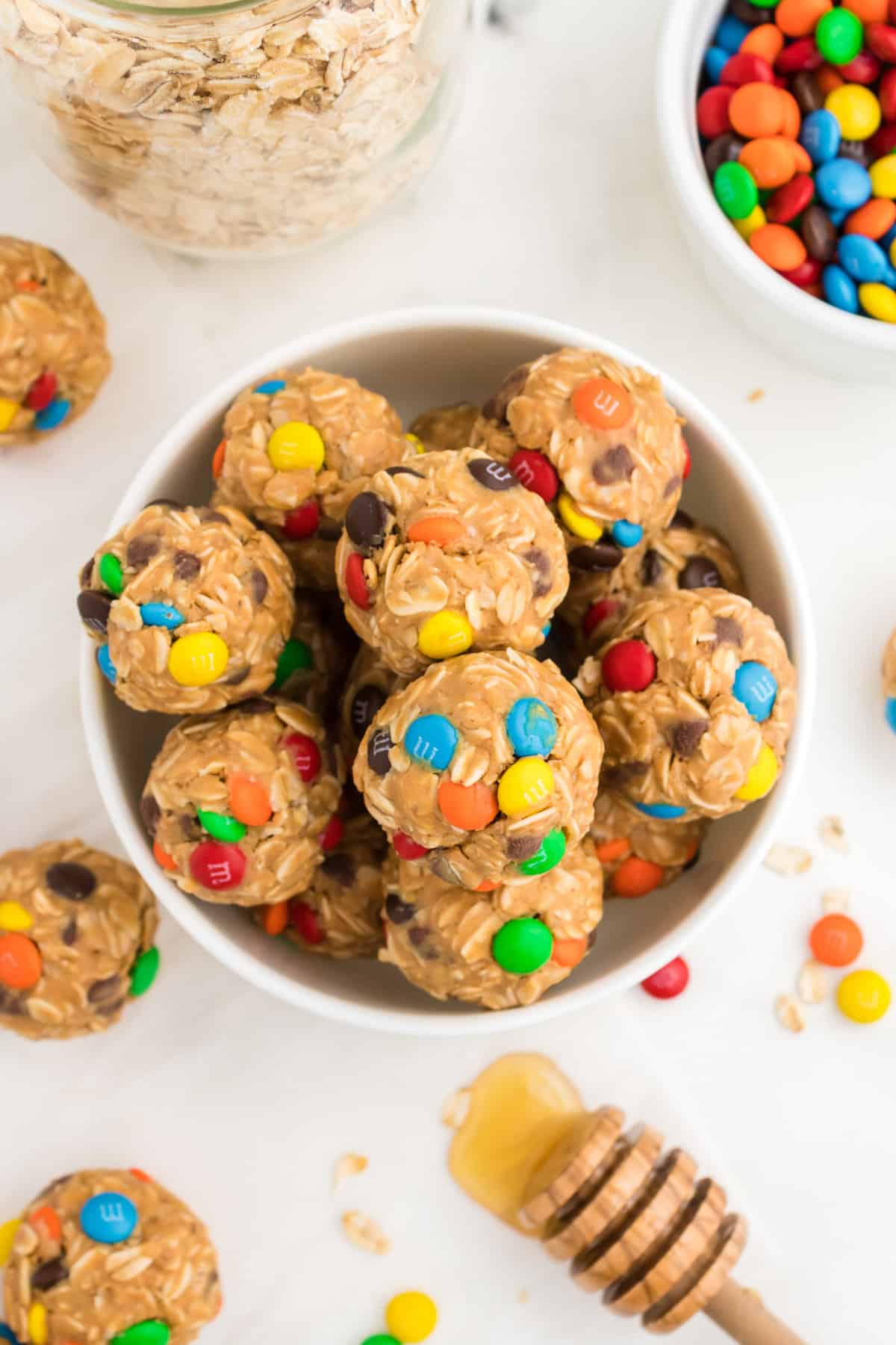 Bowl of Oatmeal Balls with M&Ms surrounded by jar of oats, honey, M&M candies, and more energy balls.