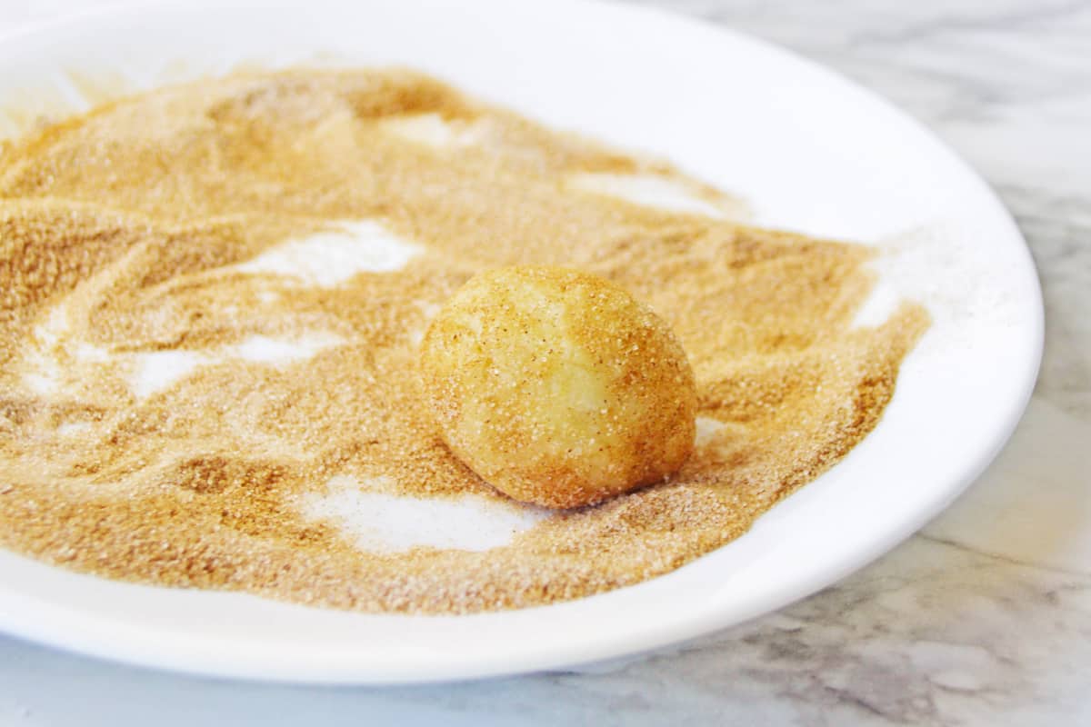 Ball of cookie dough being rolled on a plate filled with cinnamon sugar.