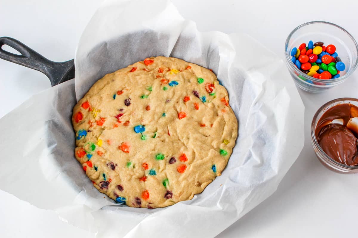 Baked skillet cookie with melted chocolate and M&Ms in bowls beside it.