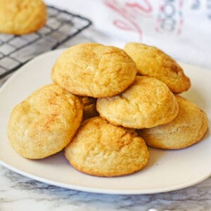 Snickerdoodle cake mix cookies stacked in a pile on a white plate with a cooling rack with additional snickerdoodles behind them.