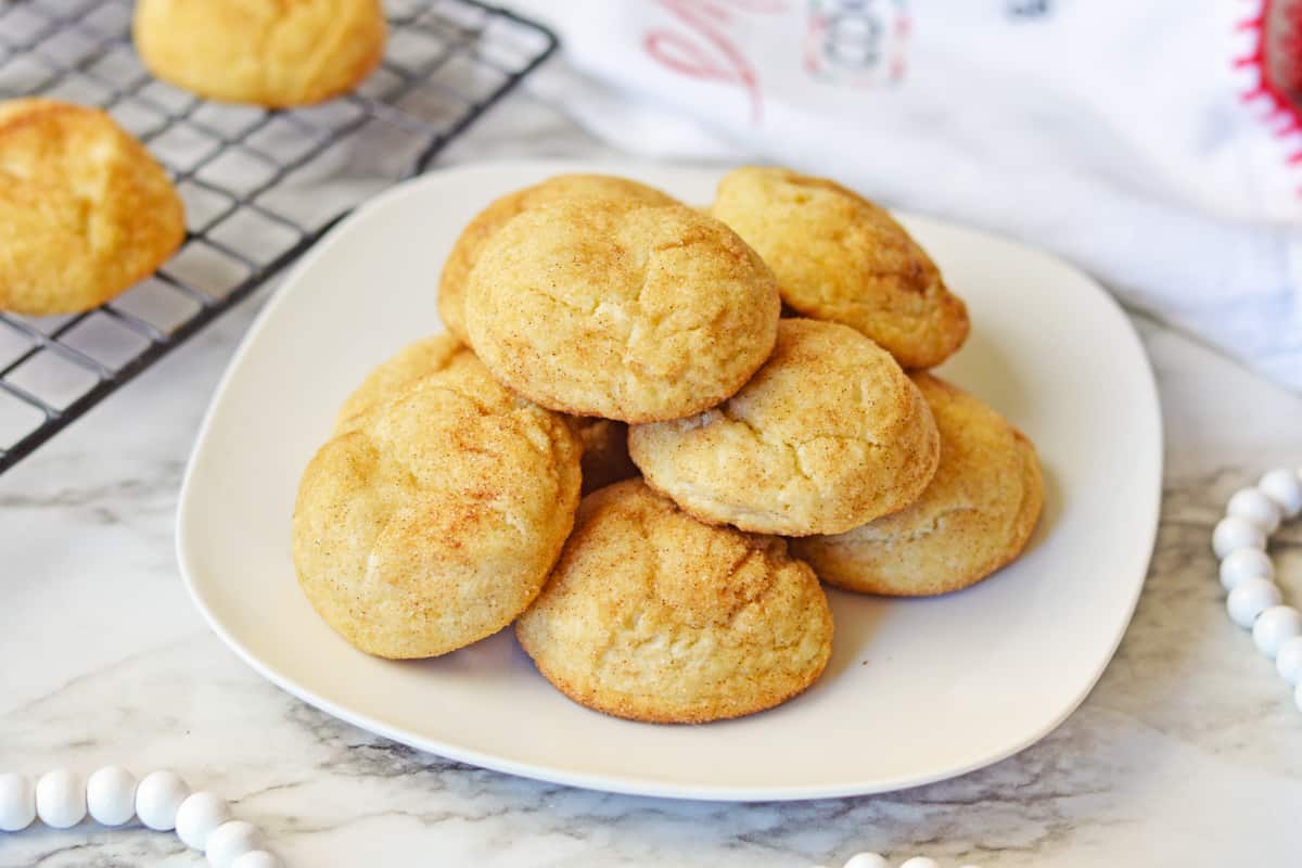 Thick snickerdoodle cake mix cookies piled on a white plate with more cookings on a cooling rack in the background.