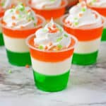 Festive St. Patrick's Day jello shots made with layers of green, white, and orange jello and topped with whipped cream and green and orange sprinkles.