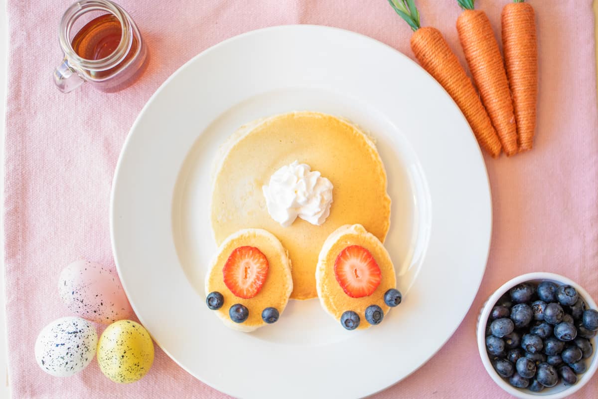 Bunny butt pancakes on white plate on pink linen with syrup, blueberries, and easter decorations around them.