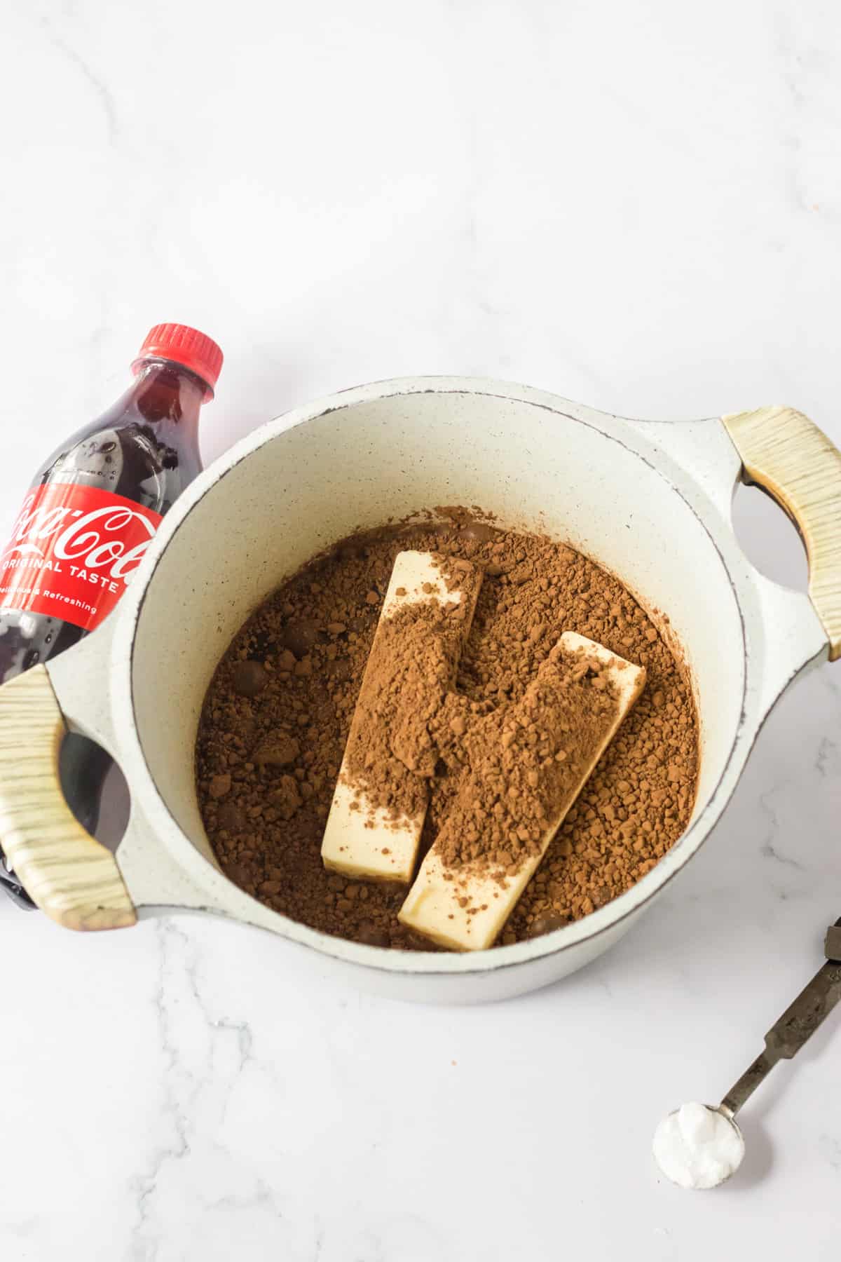 Pot with cocoa powder and two sticks of butter with a bottle of coca cola besides it.