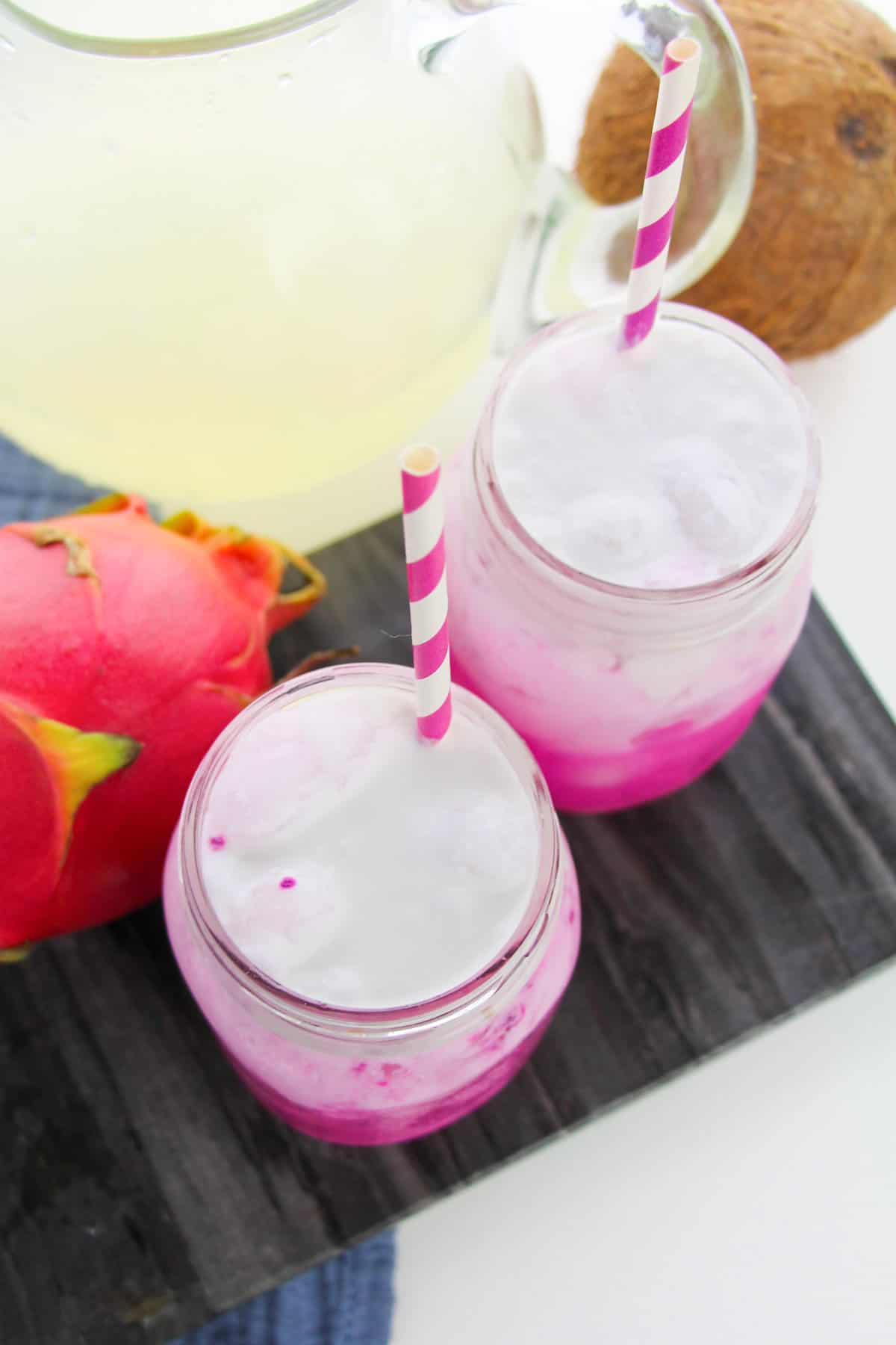 Two pink dragon drinks topped with coconut milk and whipped cream served in mason jars with pink and white striped paper straws.