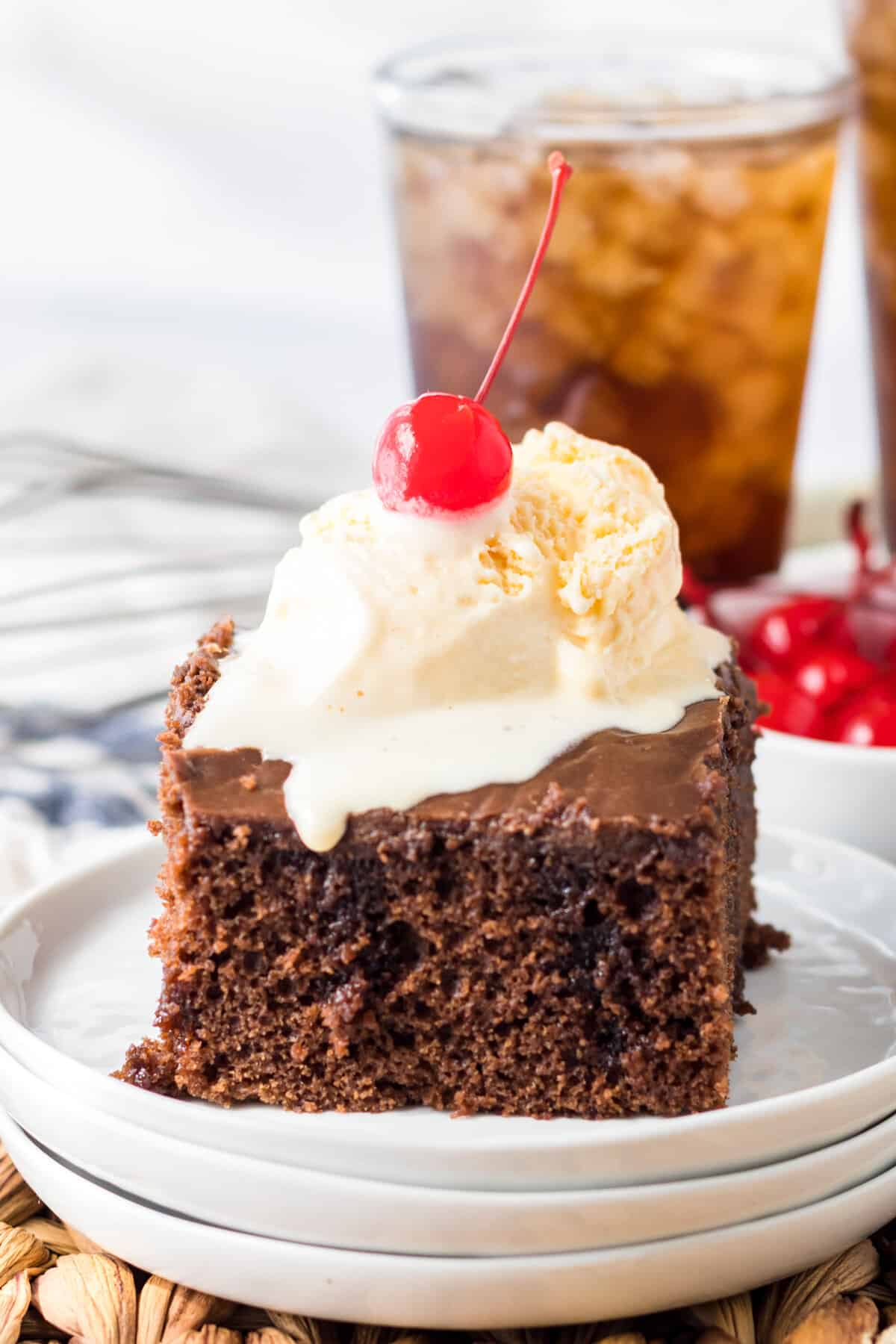 Piece of double chocolate Coca-Cola cake topped with a scoop of vanilla ice cream and a cherry. A glass of soda is behind the cake.