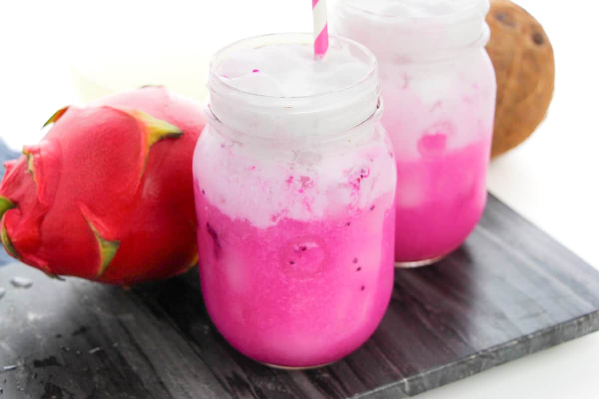 Copycat Starbucks Dragon Drink with vibrant pink mango dragonfruit refresher drink in mason jar topped with coconut milk. Another pink drink and a fresh mango and dragonfruit are in the background.