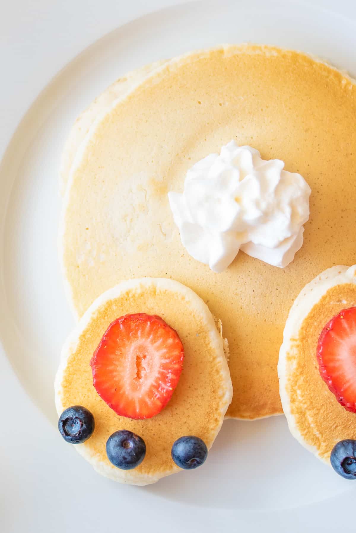 Close up of pancakes topped with strawberry slices, whipped cream, and blueberries arranged to look like an Easter bunny's butt. 