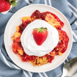 Strawberry dump cake on a white plate topped with cool whip and a fresh strawberry.