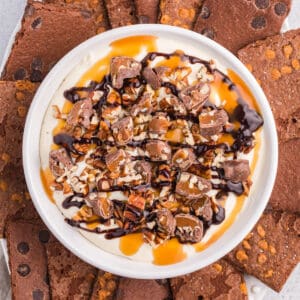 No-bake turtle cheesecake dip in a white serving bowl topped with caramel, pecans, chopped turtle candies, and served with brownie brittle for dipping.