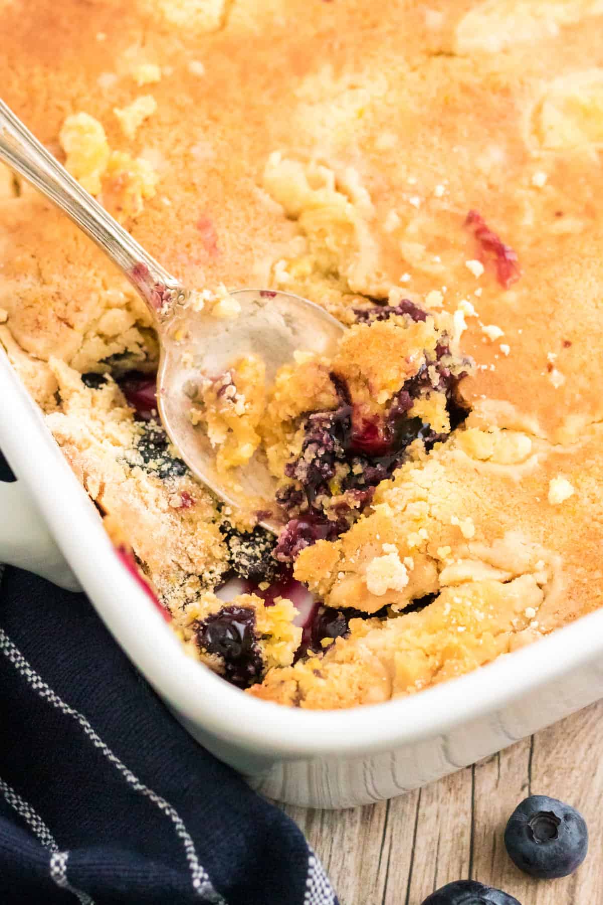 Lemon Blueberry Dump Cake in white 9 x 13 pan with serving spoon breaking through the golden cake top layer so the blueberry pie filling peeks out.