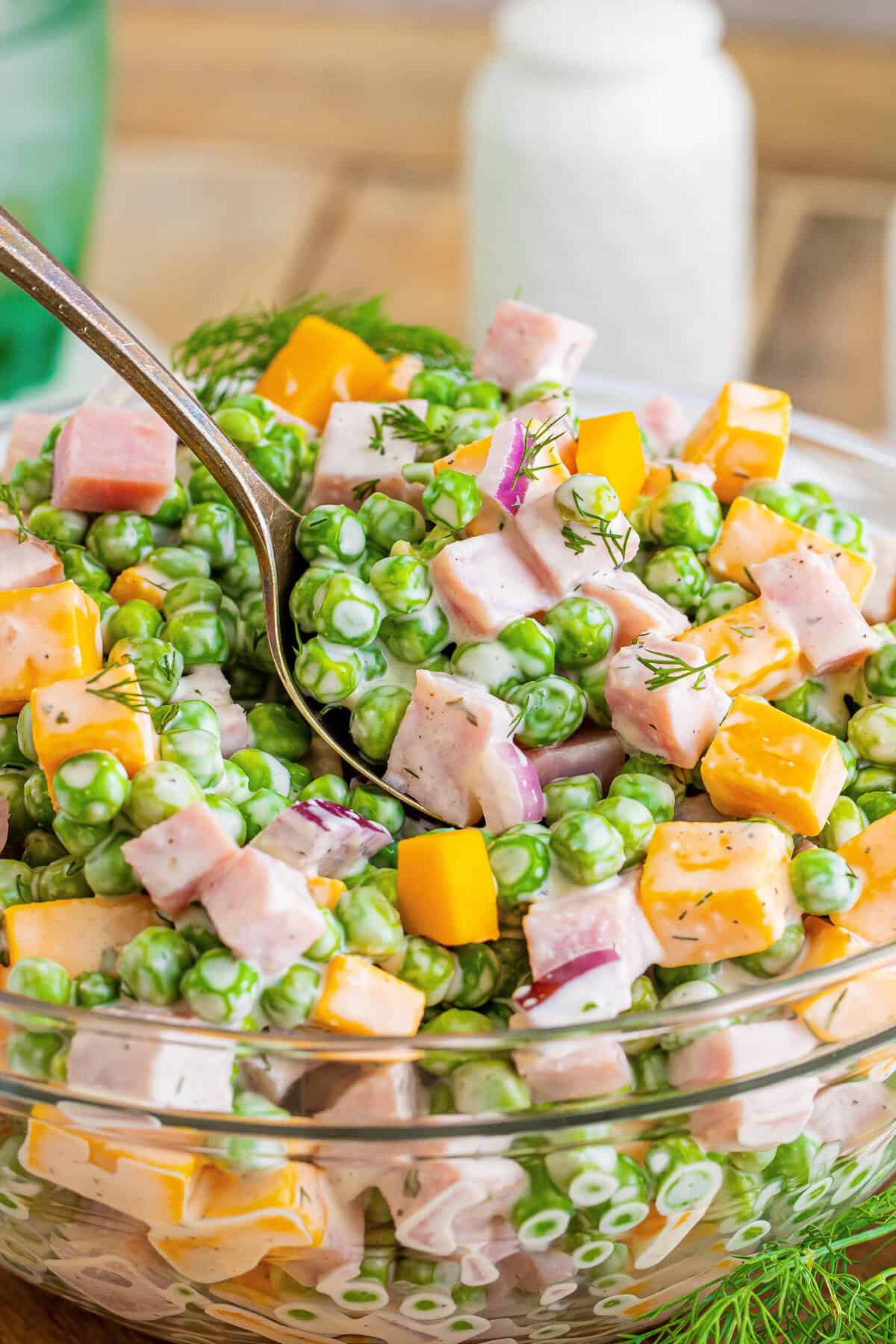 Spoon lifting a scoop of English pea salad out of large glass bowl.