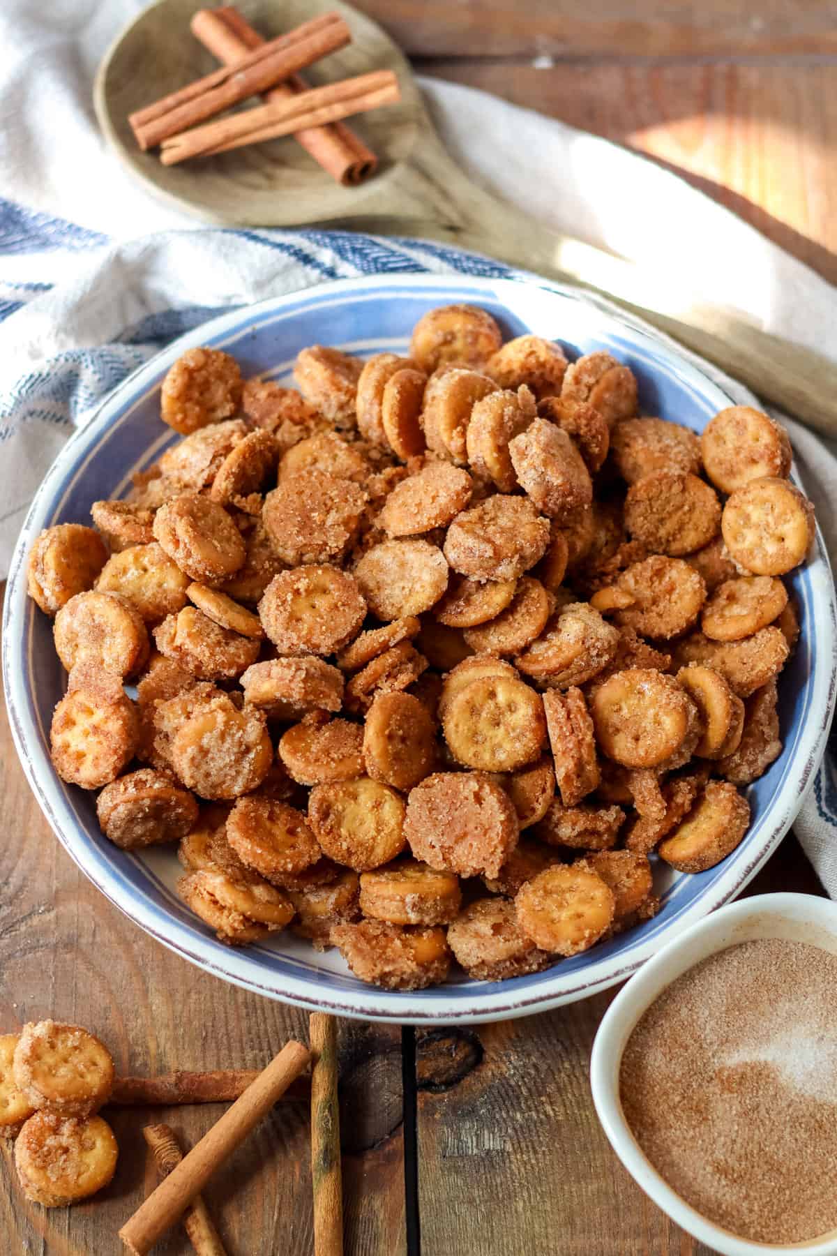 Cinnamon sugar Ritz Churro Bites in a large serving bowl on a wooden table with a small bowl of cinnamon sugar and cinnamon sticks around it.