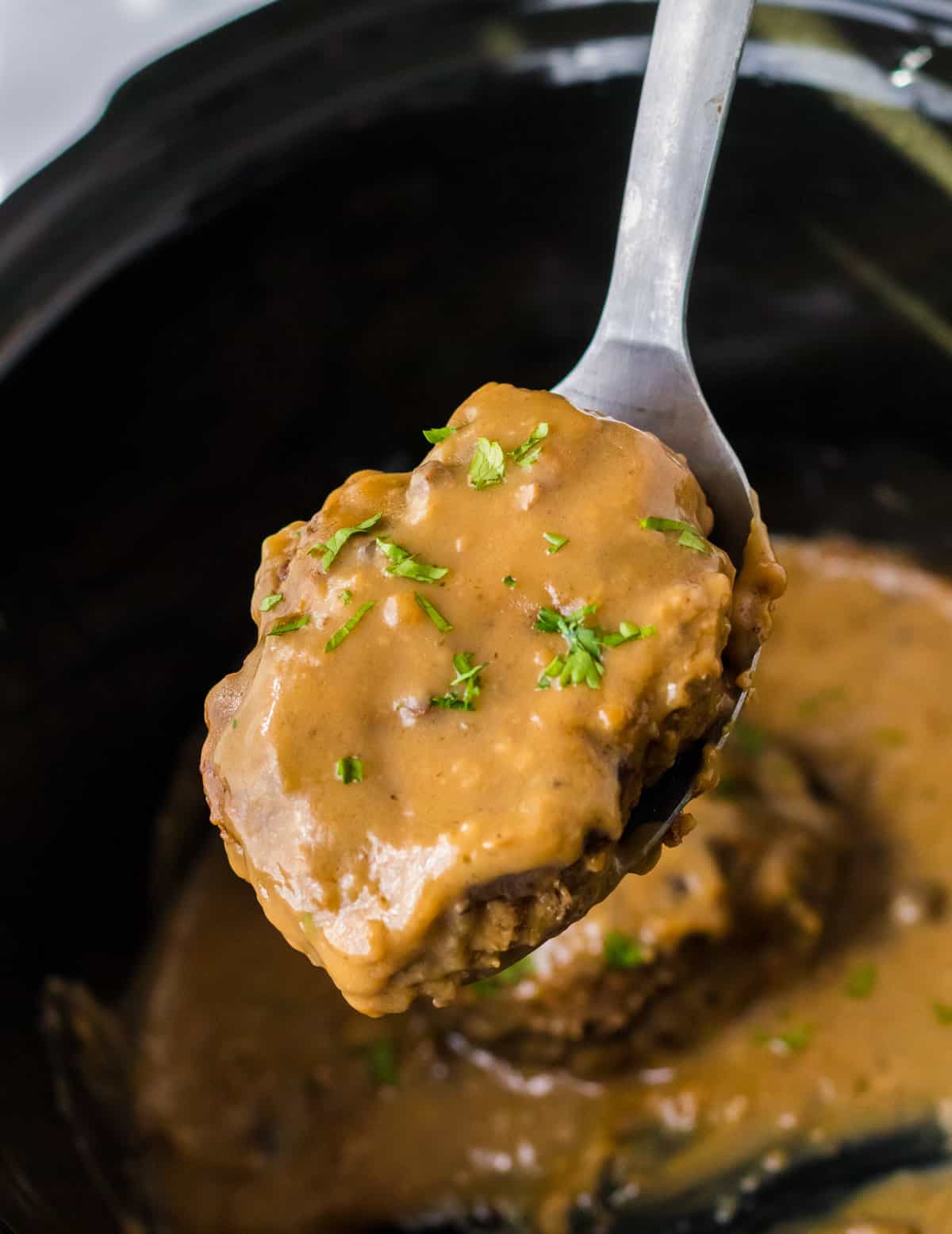 Serving spoon lifting gravy smothered salisbury steak patty out of crockpot.