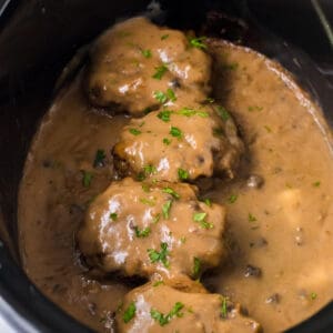 Slow cooker salisbury steaks smothered with brown gravy and garnished with parsley in a crockpot.