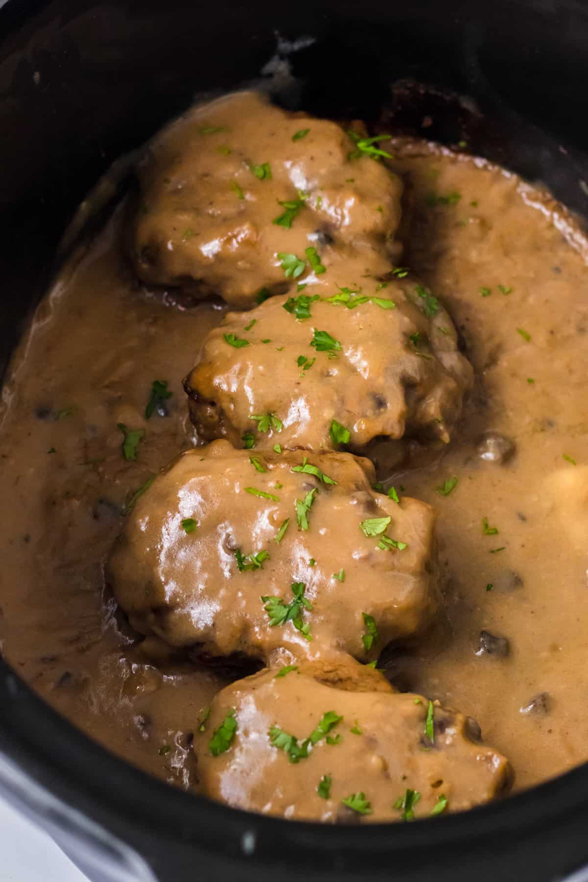 Slow cooker salisbury steaks smothered with brown gravy and garnished with parsley in a crockpot.