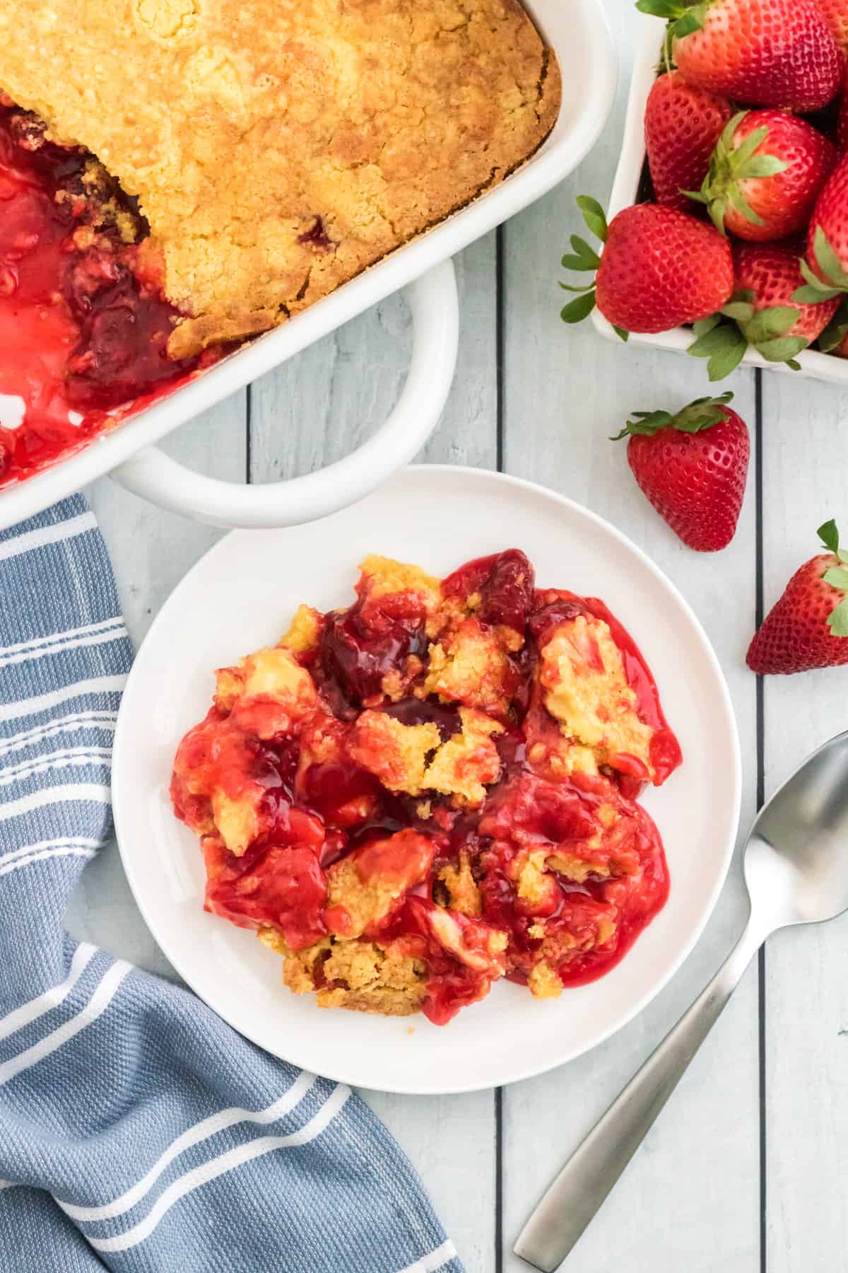 Strawberry pie filling dump cake served on a white plate with fresh strawberries and remaining cake beside it.