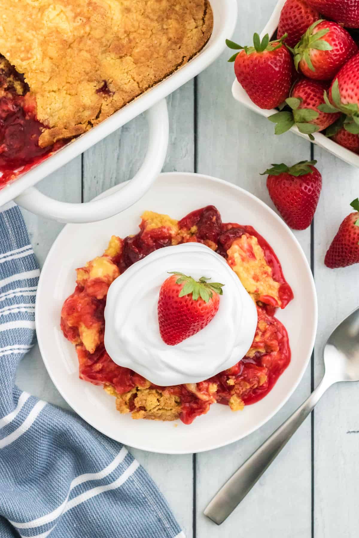 Strawberry dump cake serving on a plate topped with cool whip and a strawberry. Baking dish with remaining cake, a fork, a cloth napkin, and a pint of fresh strawberries are next to the plate.