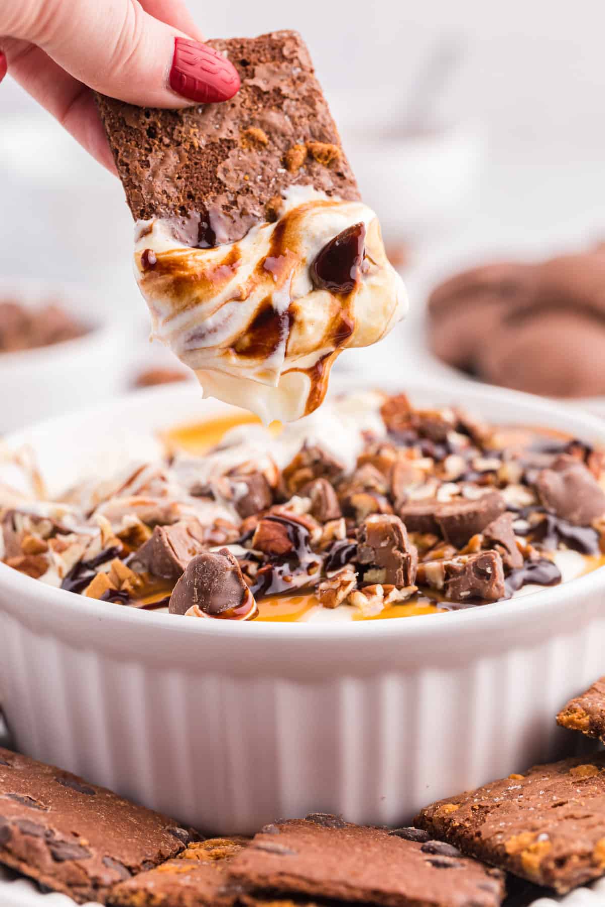 Hand reaching in to dip a chocolate cookie into the creamy no bake turtle cheesecake dip.