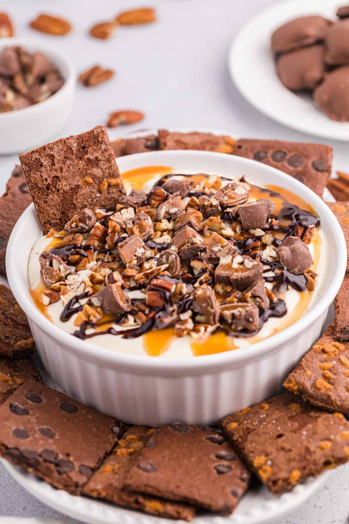 No-bake turtle cheesecake dip in a white serving bowl topped with caramel, pecans, chopped turtle candies, and served with brownie brittle for dipping.