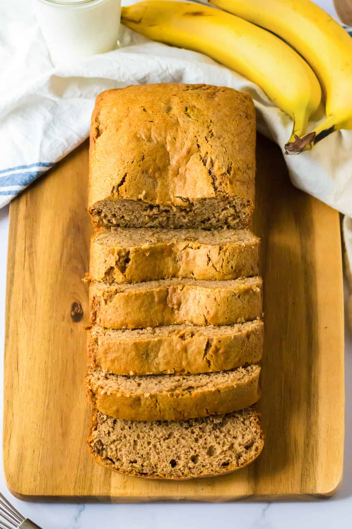 Sliced loaf of banana bread on wood cutting board with a white linen and fresh bananas.