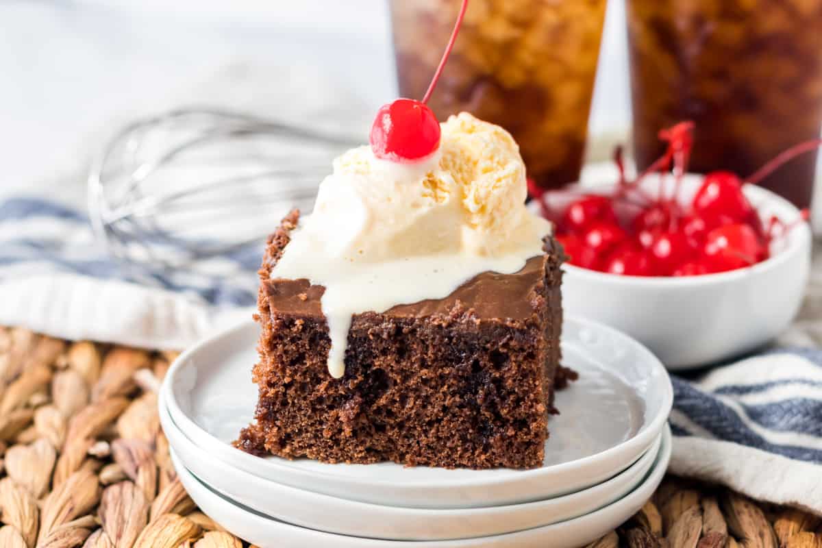 Square slice of chocolate cake with chocolate frosting and scoop of vanilla ice cream and a cherry on top.