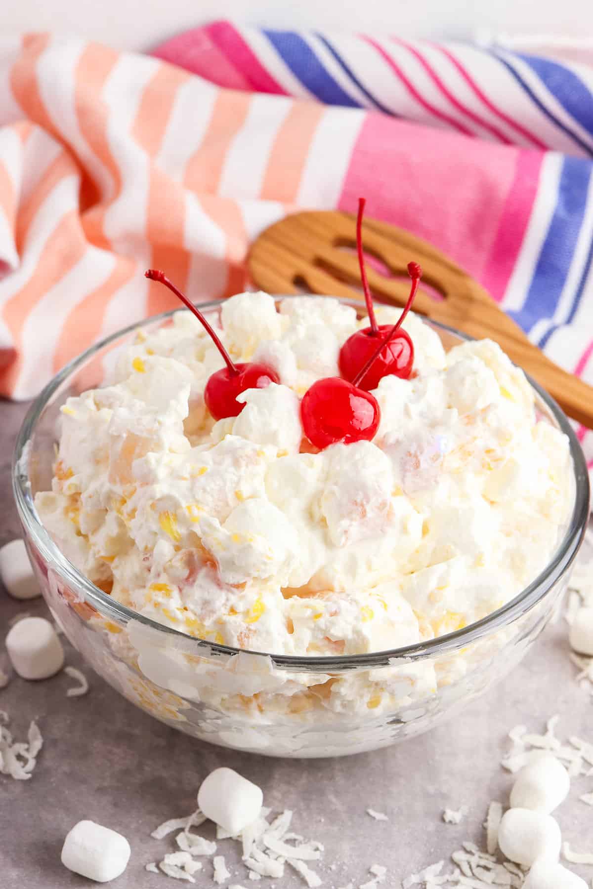 Marshmallow fruit salad with cool whip and canned fruit in a large glass bowl with colorful linen and slotted wooden spoon.