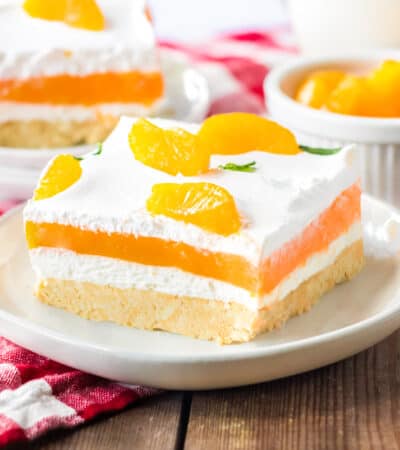 Orange creamsicle lush with layers of creamy vanilla pudding and orange jello, sweetened cream cheese, and cool whip on a sweet cookie crust. Mandarin orange pieces top the no bake cake.