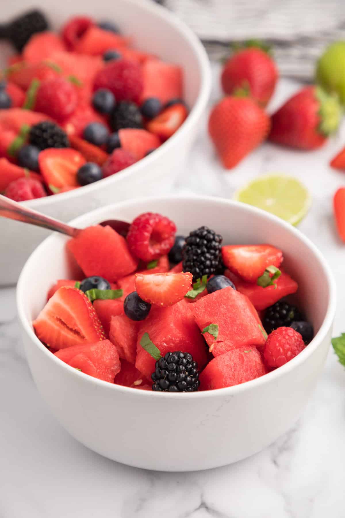 Small white bowl of watermelon fruit salad with larger serving bowl of salad in background.