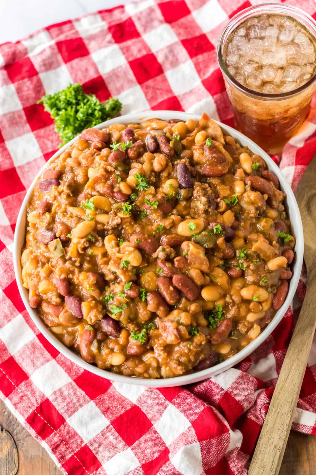 Large bowl of cowboy beans with a cup of iced tea on a red plaid tablecloth.