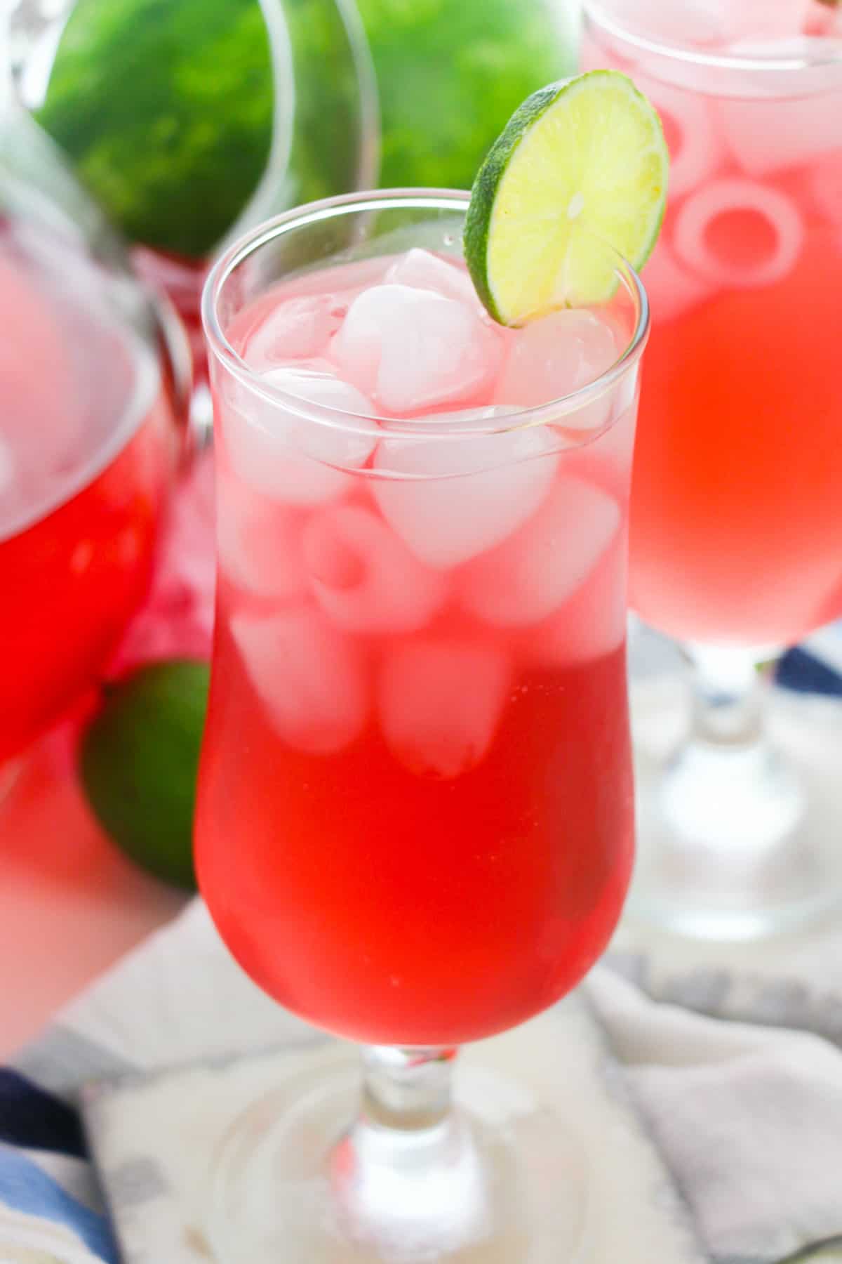 Watermelon refresher drink in glass with ice and lime wheel garnish. A pitcher and another glass of the drink are in background,