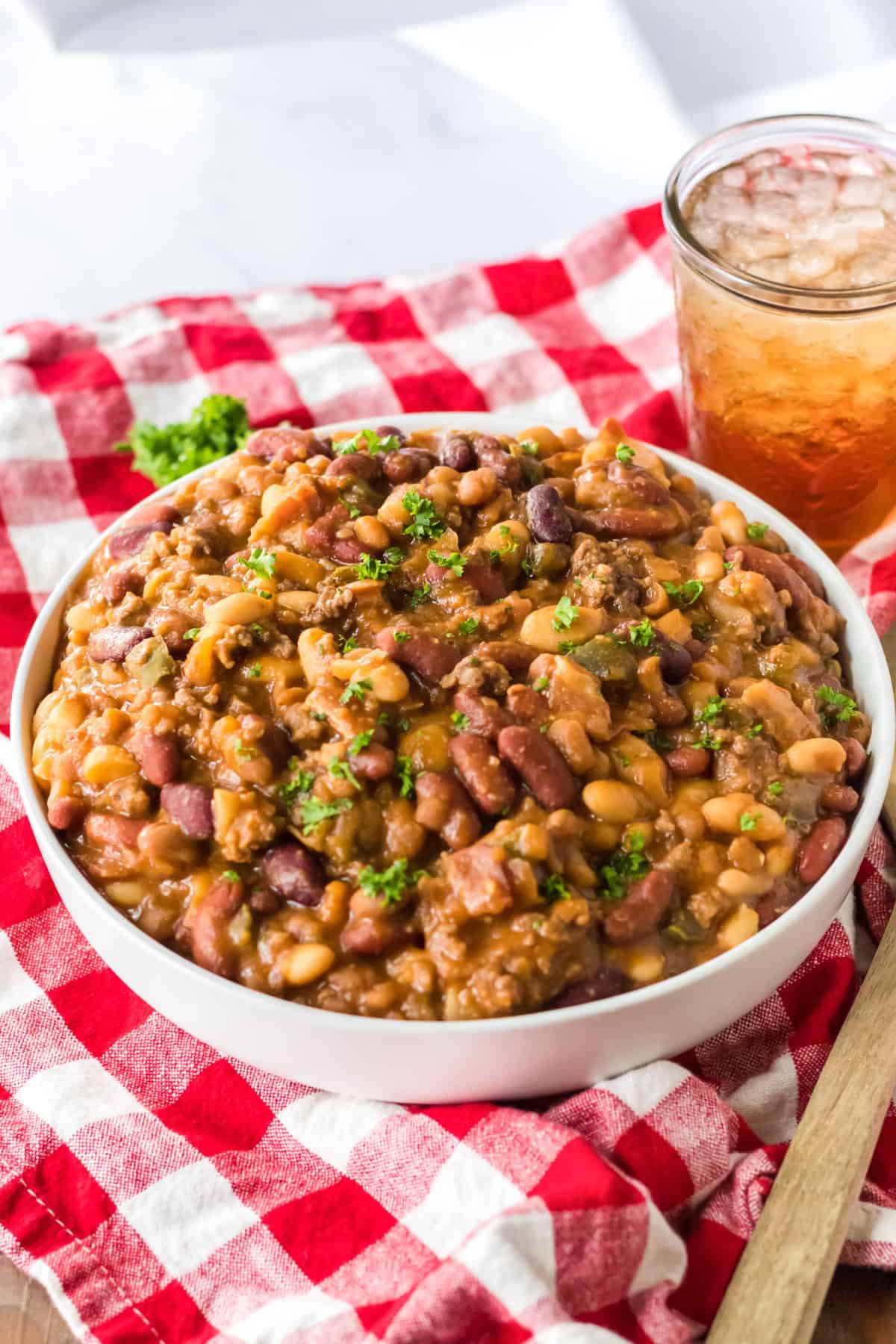 Slow cooker cowboy beans garnished with parsley and served in a large white bowl with glass of iced tea and handle of wooden spoon next to them.