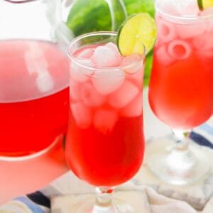 Watermelon refresher drink in glass with ice and lime wheel garnish. A pitcher and another glass of the drink are in background,